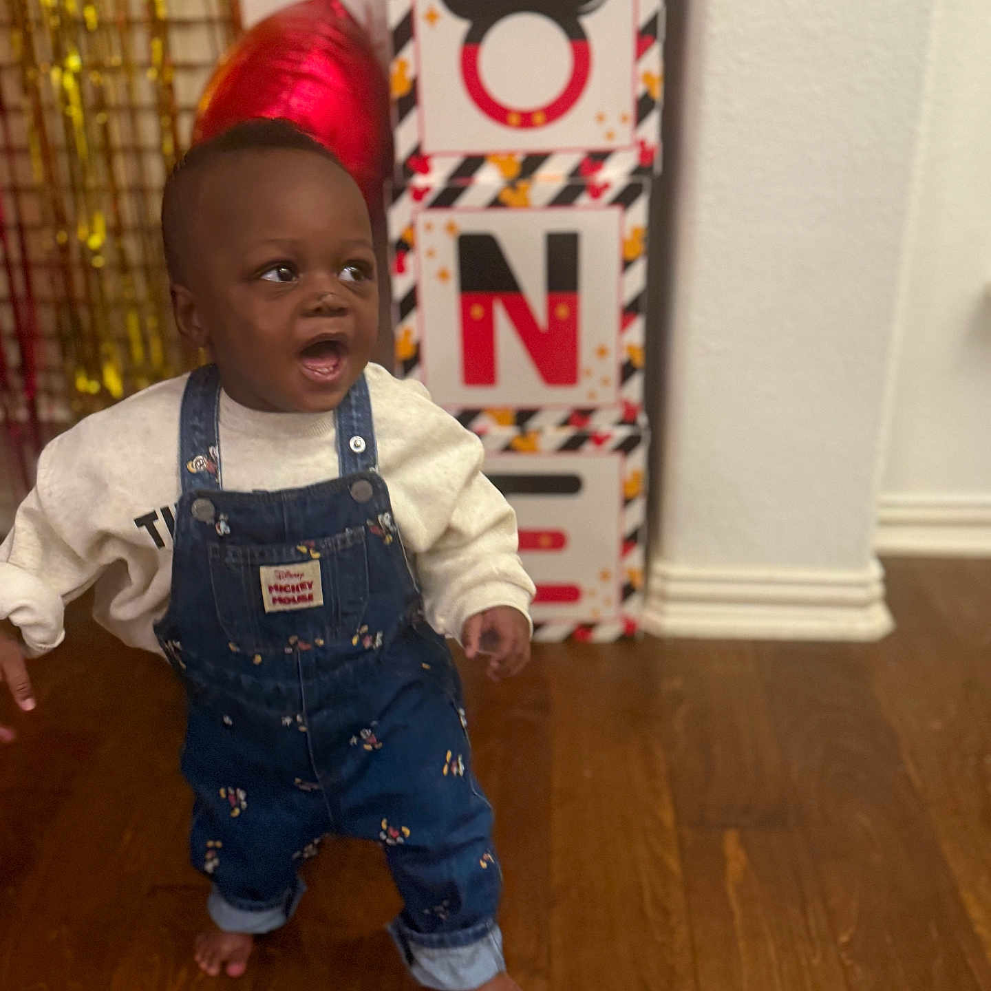 Omani joined the competition — help win amazing prizes! baby, barefoot, baseboard, celebration, child, denim_overalls, face, gift_boxes, indoor, mickey_mouse_box, party_decor, portrait, red_balloon, smile, standing, surprised_expression, sweatshirt, toddler, wall, wooden_floor