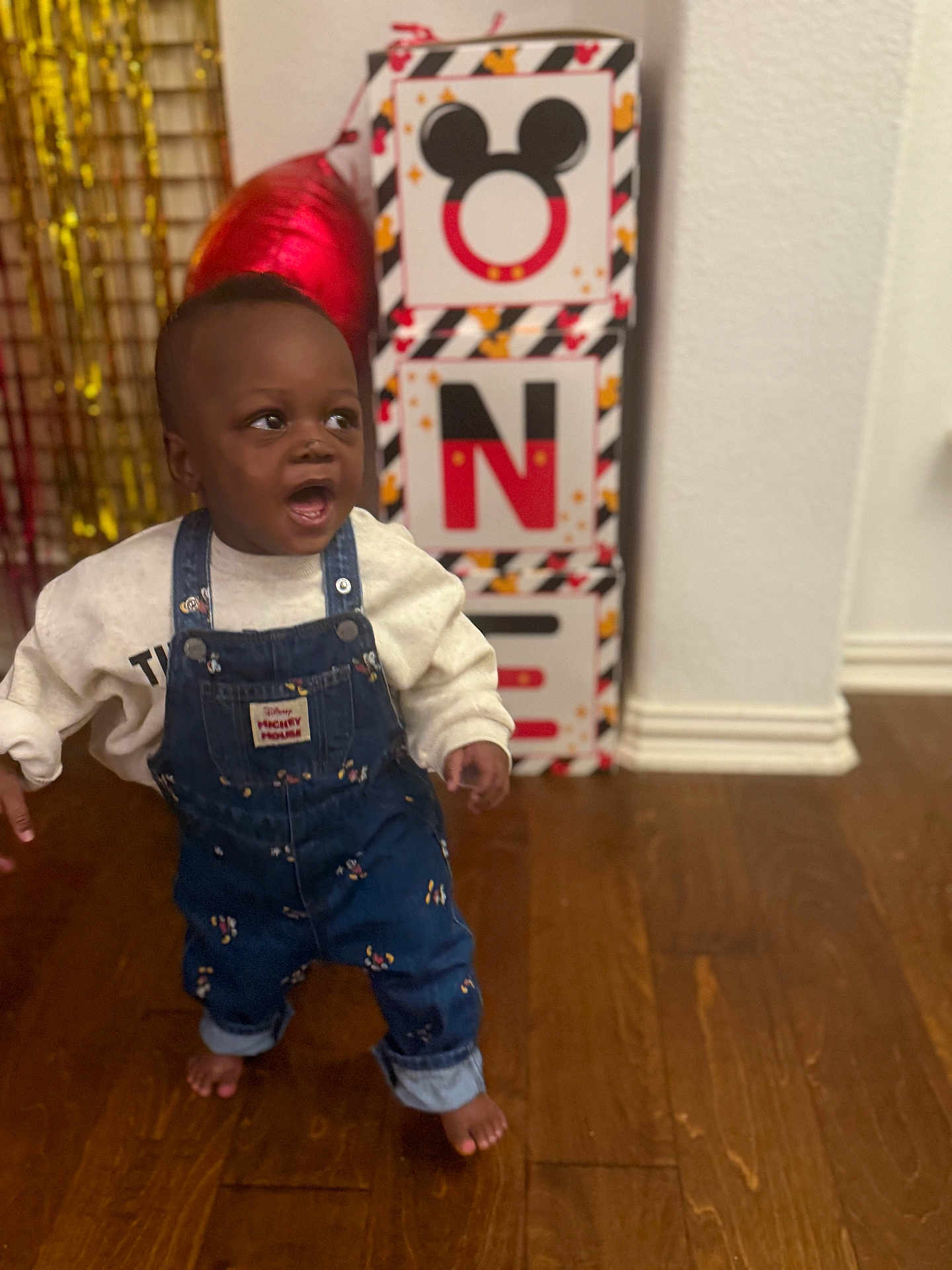Omani joined the competition — help win amazing prizes! child, toddler, baby, standing, denim_overalls, sweatshirt, barefoot, surprised_expression, mickey_mouse_box, red_balloon, party_decor, indoor, wooden_floor, baseboard, wall, gift_boxes, celebration, portrait, face, smile