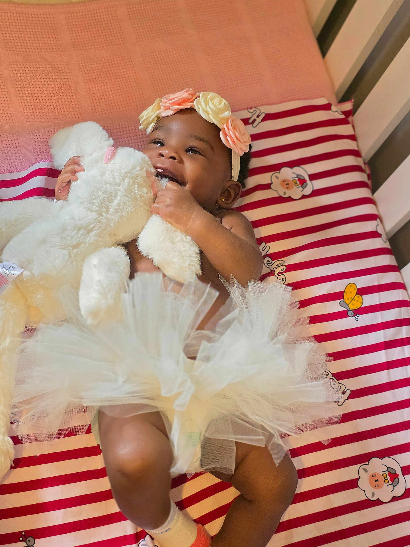 Olakamiye joined the competition — help win amazing prizes! baby, teddy_bear, headband, tutu, crib, striped_sheet, smiling, happy, toy, infant, cute, fluffy, child, indoor, soft_toy, sleeping_area, cartoon_characters, pink, white, person
