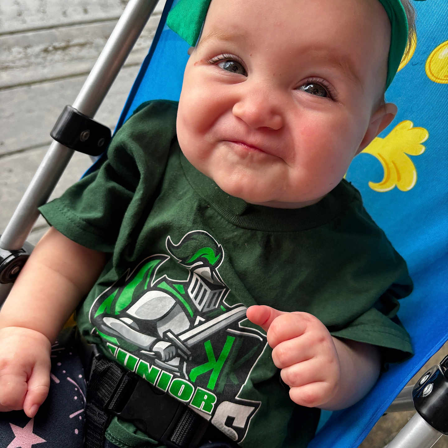 Bailey Baize is registered to the contest to win money with this photo: baby, child, infant, smiling, face, portrait, stroller, green_shirt, superhero_shirt, clothing, fist, seated, outdoor, pavement, colorful, close_up, happy, baby_hat, safety_harness, photography