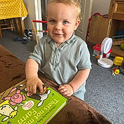 Jack joined the competition — help win amazing prizes! toddler, child, book, indoor, toy, smile, blue_eyes, blonde_hair, short_sleeve, shirt, carpet, furniture, fan, door, table, chair, wooden, colorful, playroom, happy