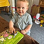 toddler, child, book, indoor, toy, smile, blue_eyes, blonde_hair, short_sleeve, shirt, carpet, furniture, fan, door, table, chair, wooden, colorful, playroom, happy