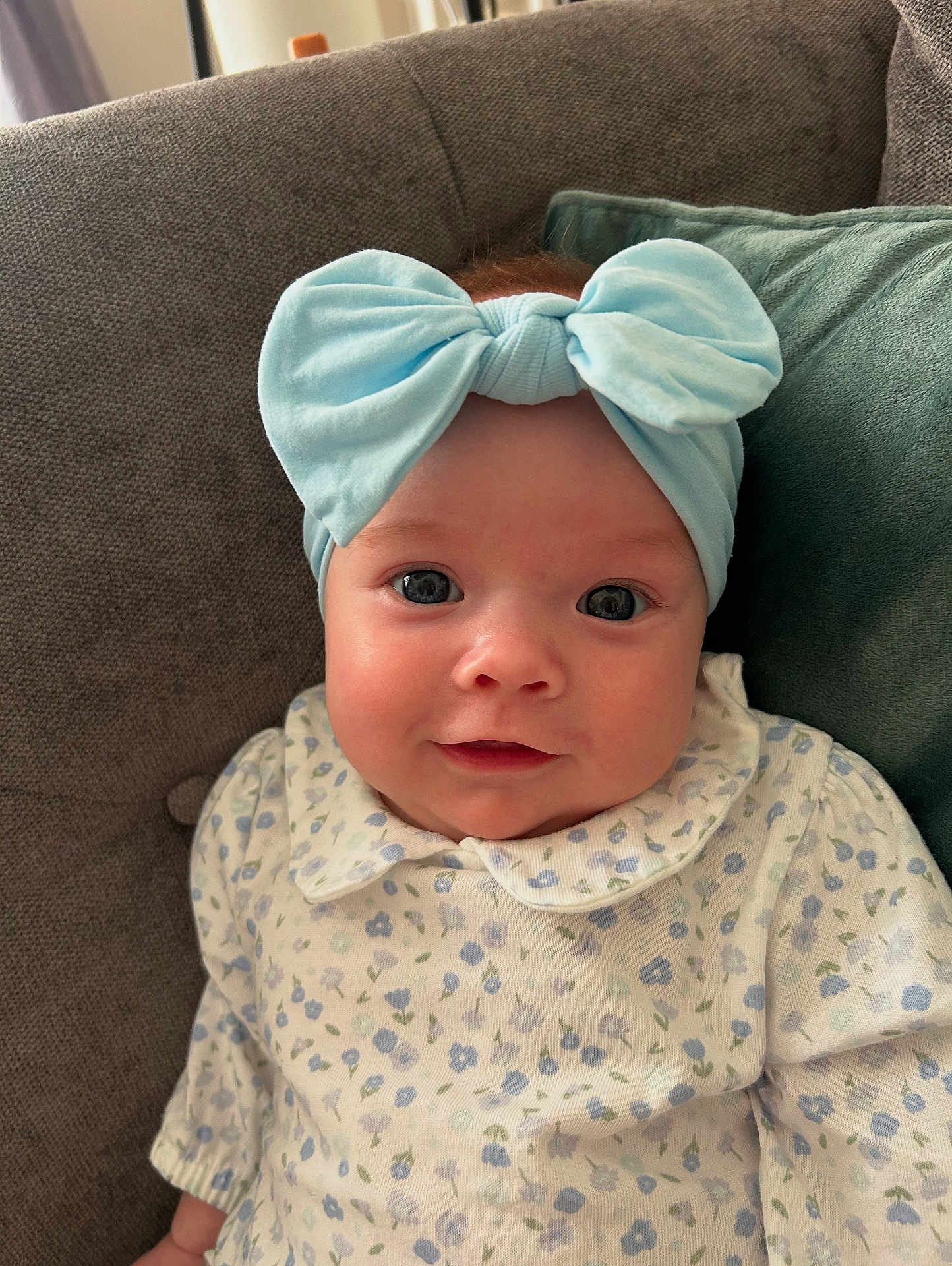 Clodagh joined the competition — help win amazing prizes! baby, child, infant, headband, bow, smile, face, eyes, couch, pillow, clothing, pattern, floral, sitting, indoors, portrait, cute, young, soft_lighting, happy