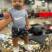 Manuel is registered to the contest to win money with this photo: toddler, child, baby, pacifier, blue_onesie, nike_logo, shorts, white_sneakers, hand_support, camo_tablecloth, indoor, market, blurred_background, person, footwear, clothing, cute, standing, infant, fashion