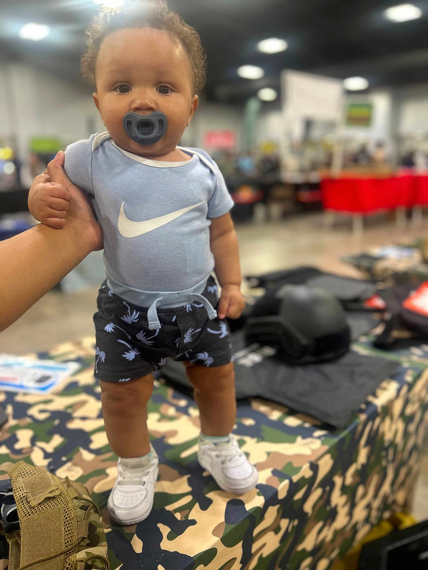 Manuel is registered to the contest to win money with this photo: toddler, child, baby, pacifier, blue_onesie, nike_logo, shorts, white_sneakers, hand_support, camo_tablecloth, indoor, market, blurred_background, person, footwear, clothing, cute, standing, infant, fashion