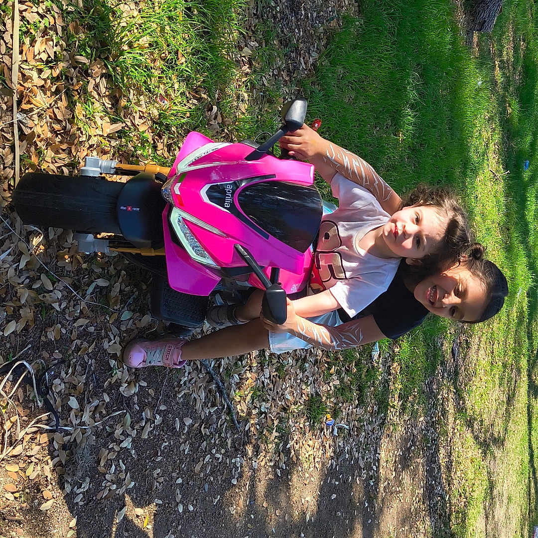 Atreus is registered to the contest to win money with this photo: children, toy_motorcycle, pink, outdoor, grass, leaves, sunlight, shadow, play, happiness, fun, park, casual_clothing, smile, nature, dirt_path, sneakers, summer, friendship, child