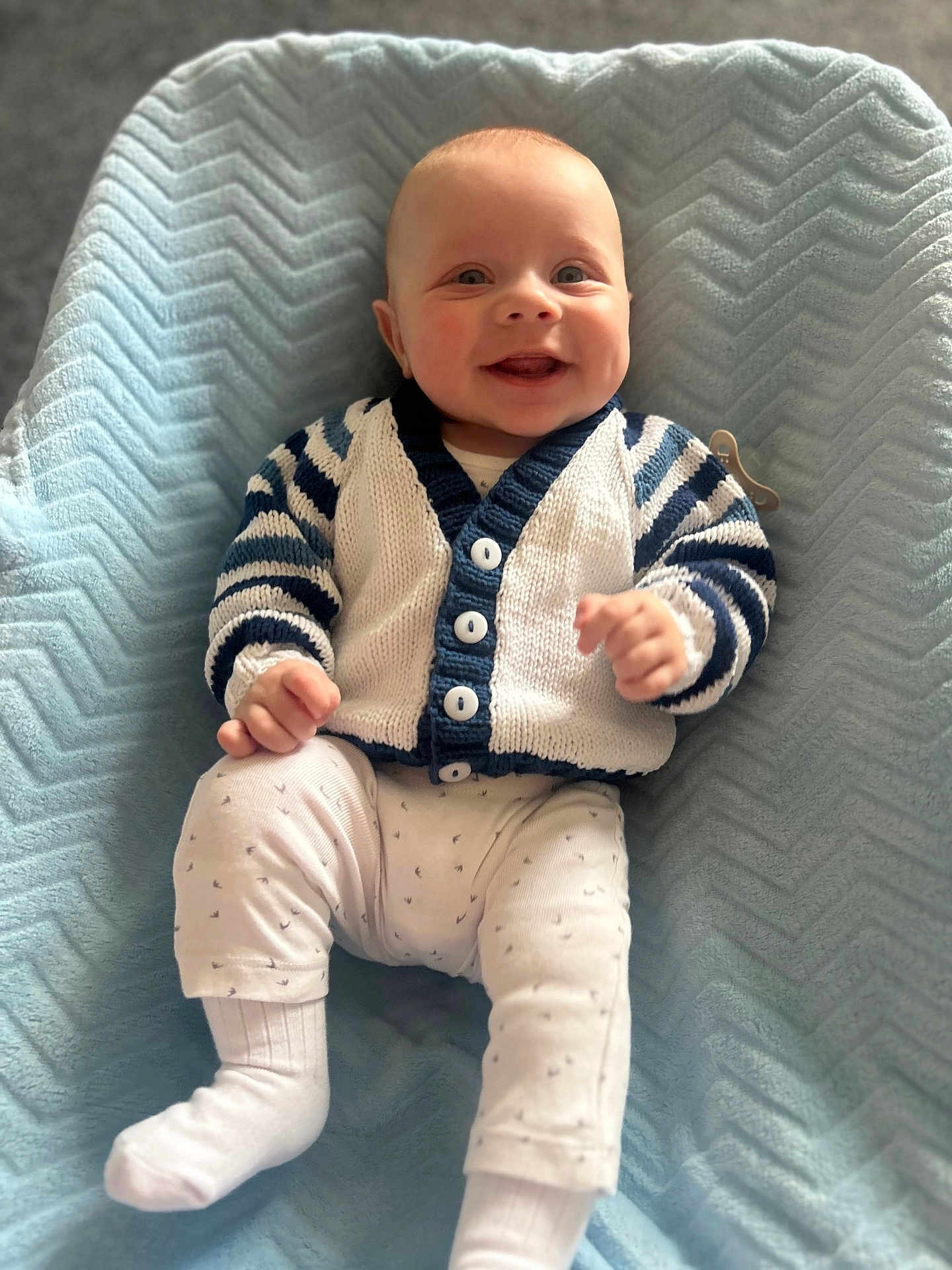 Corey is registered to the contest to win money with this photo: baby, smiling, child, sitting, blanket, blue, cardigan, striped, white_pants, socks, happy, portrait, indoor, cozy, cute, infant, soft_texture, knitted_clothing, face, person