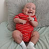 baby, bed, child, chubby_legs, cute, face, happy, indoor, infant, joyful, person, pillow, playful, plush_pillow, red_onesie, sitting, smiling, soft_texture, striped_clothing, white_socks