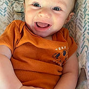 Noah is registered to the contest to win money with this photo: baby, child, smiling, orange_shirt, hands, face, cute, infant, happy, blue_eyes, patterned_background, cushion, lying_down, skin, portrait, closeup, young, person, indoors, expression