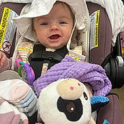Ava is registered to the contest to win money with this photo: baby, infant, smiling, hat, car_seat, seat_belt, plush_toy, blanket, teether, socks, clothing, happy, close_up, portrait, store, warning_label, strap, toy, soft_toy, purple_blanket