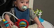 Aaron joined the competition — help win amazing prizes! baby, child, infant, pacifier, activity_center, jumper, toy, colorful, sitting, indoors, portrait, wide_eyed, brown_hair, bib, blue_clothing, gray_shirt, playroom, furniture, happy, photograph