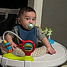 baby, child, infant, pacifier, activity_center, jumper, toy, colorful, sitting, indoors, portrait, wide_eyed, brown_hair, bib, blue_clothing, gray_shirt, playroom, furniture, happy, photograph