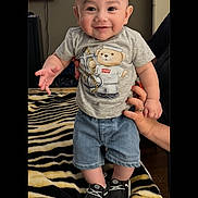 Aaron is registered to the contest to win money with this photo: baby, child, standing, smiling, happy, portrait, indoors, face, person, infant, clothing, t_shirt, shorts, sneakers, blanket, zebra_pattern, adult_hand, furniture, smartphone_screenshot, notification