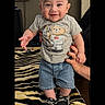 baby, child, standing, smiling, happy, portrait, indoors, face, person, infant, clothing, t_shirt, shorts, sneakers, blanket, zebra_pattern, adult_hand, furniture, smartphone_screenshot, notification