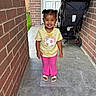 brick_wall, child, curly_hair, daylight, doorway, family_photo, front_yard, happy, outdoor, patio, paved_floor, pink_pants, portrait, sandals, smiling, smiling_face, soccer_shirt, standing, stroller, toddler