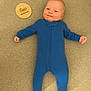 baby, infant, blue_onesie, smiling, lying_down, milestone_marker, wooden_marker, two_months, soft_surface, beige_background, cute, child, newborn, portrait, happy, small_hands, cozy_clothing, baby_clothes, young_baby, face