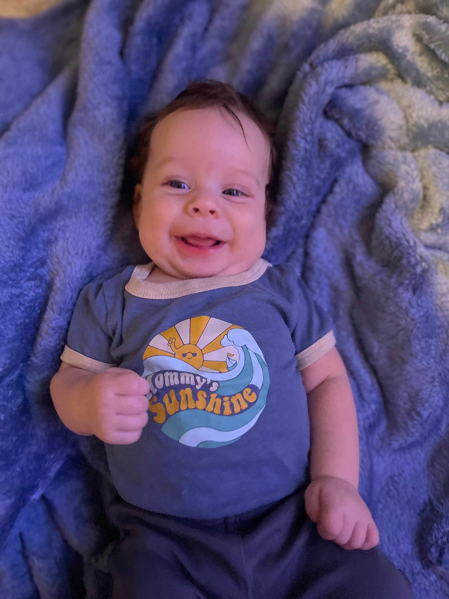 Silas is registered to the contest to win money with this photo: baby, smiling, blue_blanket, tshirt, text_on_clothing, infant, happy, lying_down, cute, cheerful, face, person, clothing, young_child, indoors, soft_texture, skin, hand, portrait, head