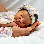 Lina is registered to the contest to win money with this photo: newborn, baby, infant, sleeping, hand, floral_pattern, headband, soft_blanket, close_up, portrait, tenderness, care, resting, peaceful, human_skin, adult_hand, white_background, wrapped, cute, child