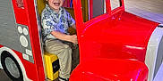 Jaxon is registered to the contest to win money with this photo: background, boy, child, clothing, entertainment, fire_truck, floor, footwear, fun, happy, indoor, light, pants, playful, ride, seat, short_sleeve_shirt, smiling, toy, vehicle