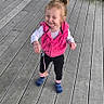toddler, child, smiling, standing, wooden_deck, pink_vest, black_pants, blue_sock, pink_sock, outdoor, person, happy, casual_clothing, young_child, hair_tied, feet, playful, daylight, cute, fun
