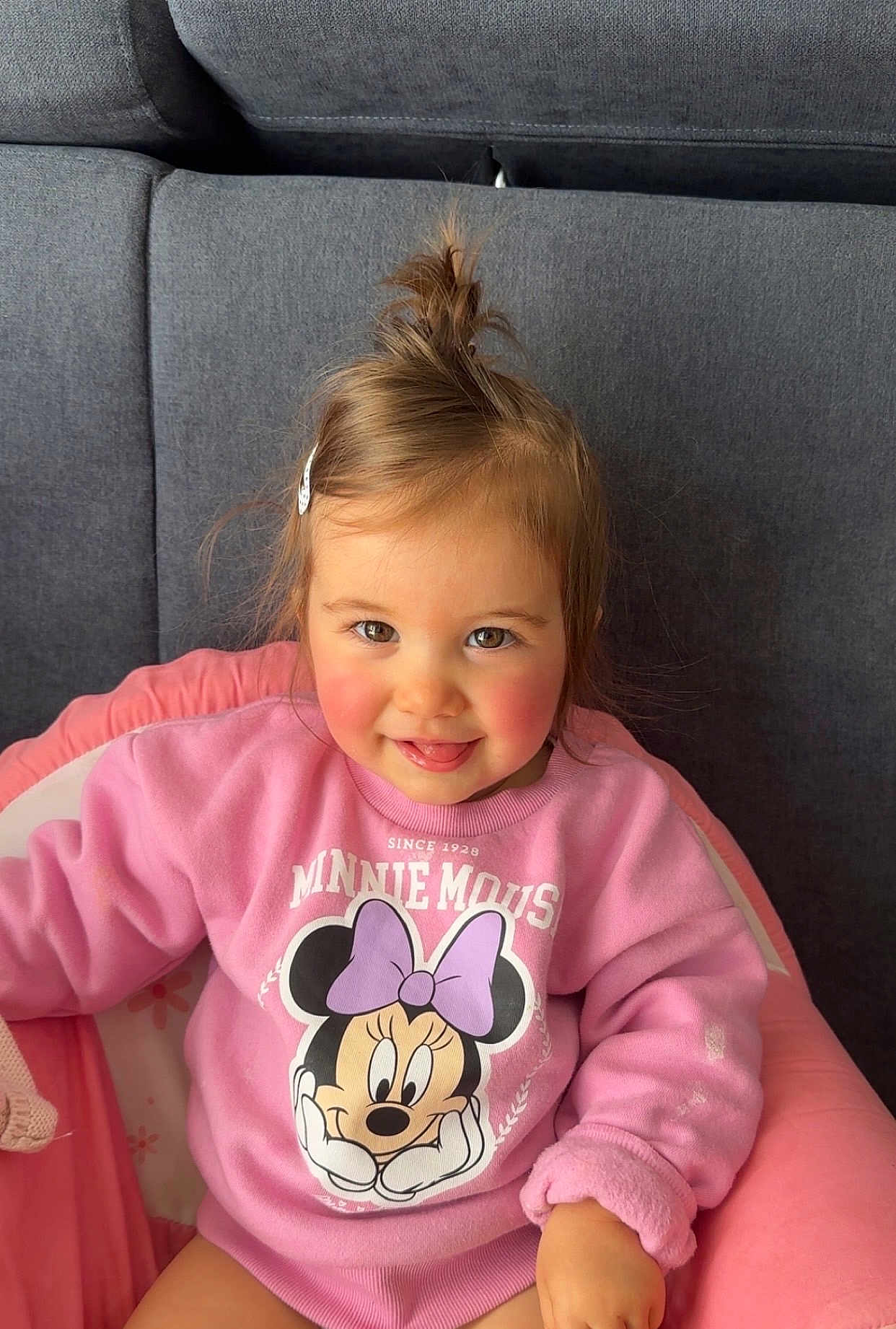Leiya participe au concours pour gagner de l'argent avec cette photo : child, toddler, girl, smiling, tongue_out, pink_sweatshirt, minnie_mouse, bow_graphic, hair_clip, top_knot, rosy_cheeks, couch, baby_chair, portrait, indoor, clothing, adorable, brown_hair, eyes, hands
