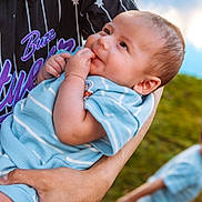 Zaiel is registered to the contest to win money with this photo: baby, child, infant, toddler, outdoor, portrait, baseball_jersey, blue_clothing, adult, arms, smiling, looking_at_camera, hand, chewing, grass, sky, clouds, blurred_background, family, daylight