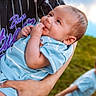 baby, child, infant, toddler, outdoor, portrait, baseball_jersey, blue_clothing, adult, arms, smiling, looking_at_camera, hand, chewing, grass, sky, clouds, blurred_background, family, daylight