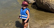 Naveen is registered to the contest to win money with this photo: child, boy, outdoor, lake, water, rocks, boulders, life_jacket, cap, smiling, standing, swimming, shoreline, bucket, water_toy, sunlight, nature, adventure, reflection, portrait