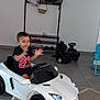 bag, black_shirt, black_wheels, car_headlights, carpet, child, clapping, floor_tiles, happy, home_interior, indoor, person, play, red_number, shoe_rack, shoes, toddler, toy_car, white_wall, young_child