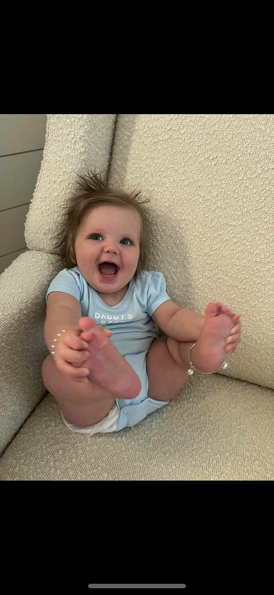 Boston is registered to the contest to win money with this photo: baby, infant, smiling, happy, blue_onesie, barefoot, armchair, beige, textured_fabric, indoor, cute, child, person, playful, hair, skin, foot, hand, sitting, joy