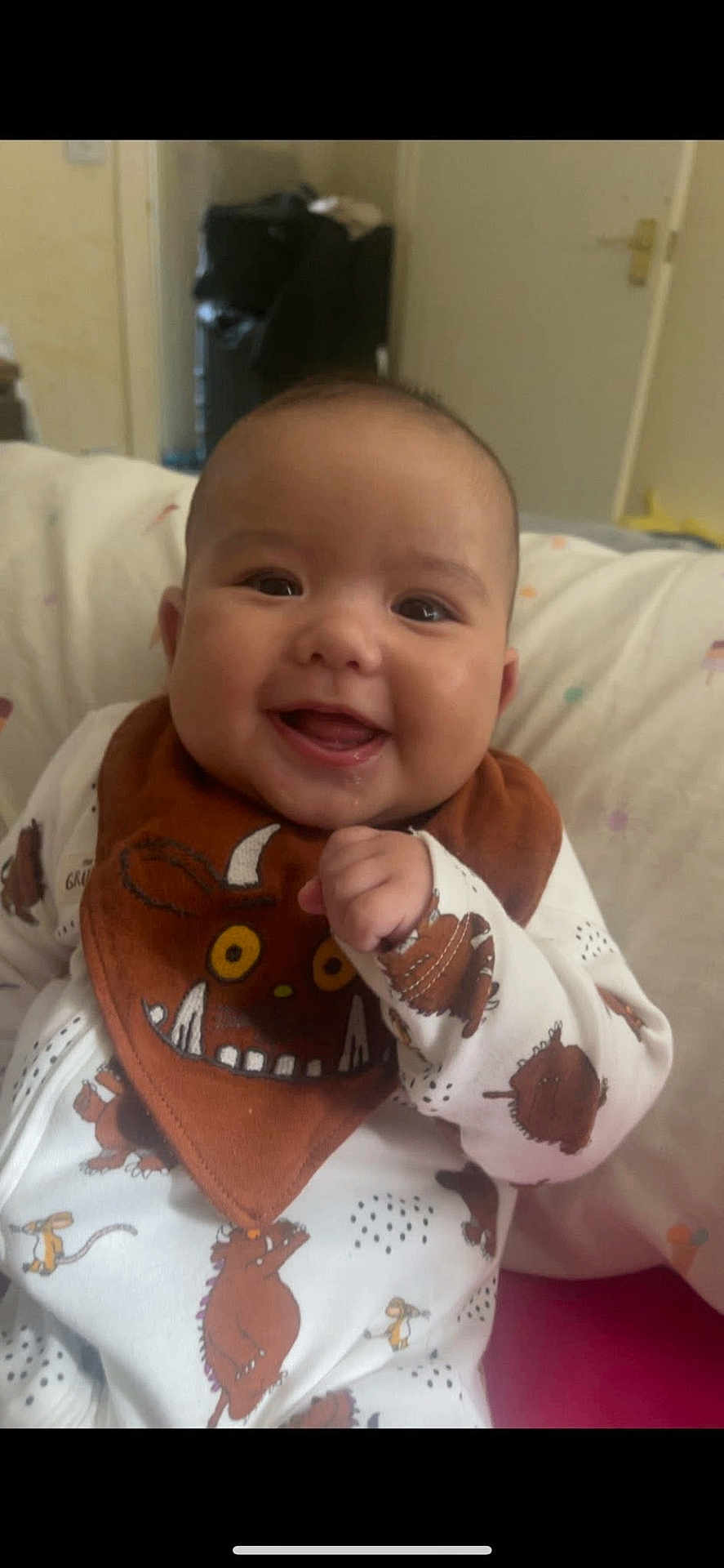 Noah is registered to the contest to win money with this photo: baby, smiling, infant, bib, clothing, cute, face, indoors, pillow, sitting, child, young, portrait, happy, white_outfit, animal_print, cozy, cheerful, hand, expression