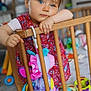 background_blur, blue_eyes, child, clothing, colorful, crib, curious, cute, flooring, hair_buns, home, indoor, leaning, person, playroom, red_dress, soft_lighting, toddler, toy, wooden_crib