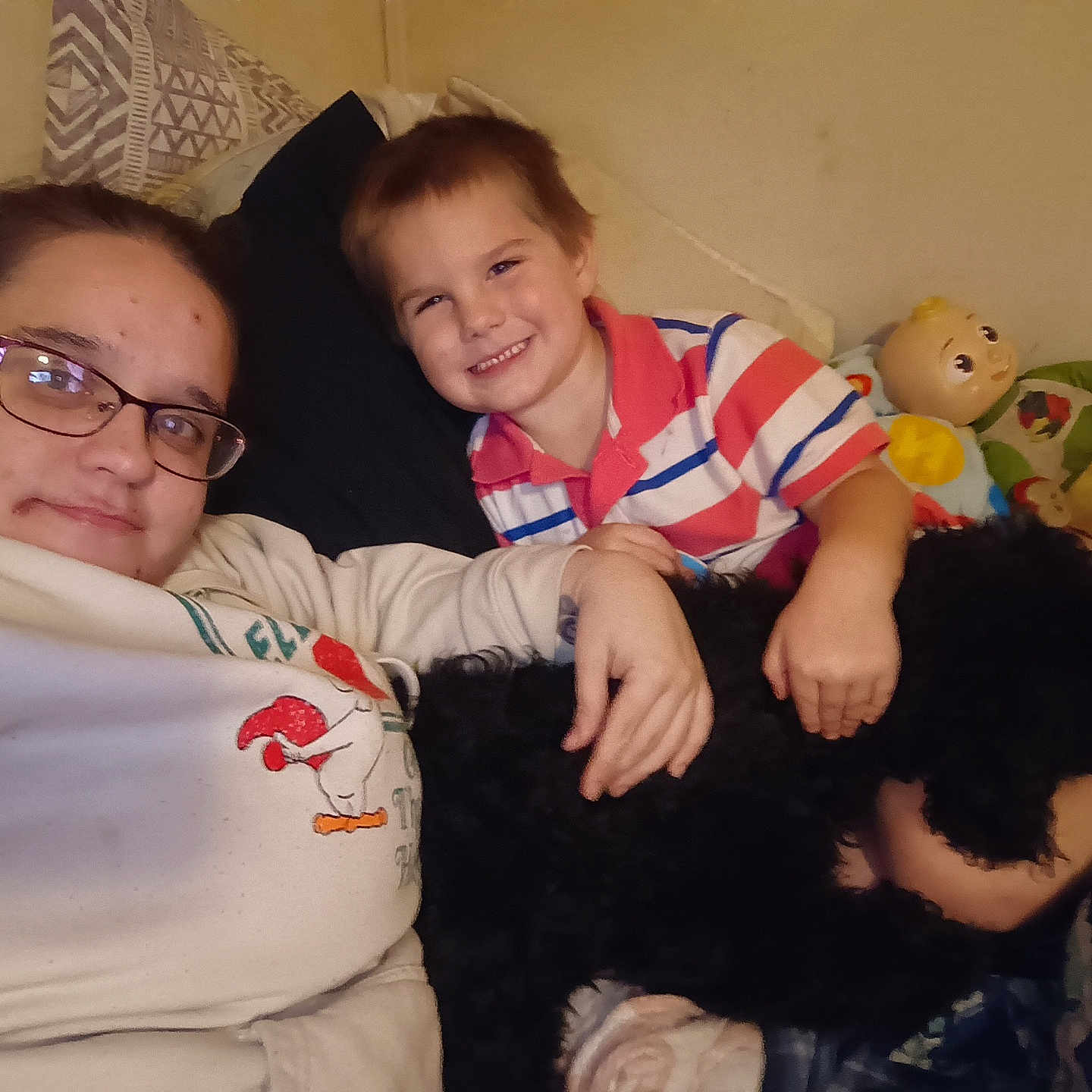 Levie is registered to the contest to win money with this photo: adult, bed, black_dog, blanket, casual_clothing, child, cuddling, dog, family, glasses, happy, home, indoor, person, pillow, portrait, relaxed, smile, striped_shirt, toy