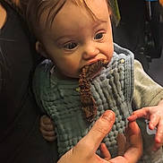Carolynn is registered to the contest to win money with this photo: adult_hand, baby, baby_carrier, bib, child, closeup, clothing, curious, feeding, food, hand, human, indoor, infant, messy_face, person, portrait, seat, wide_eyes, young_child