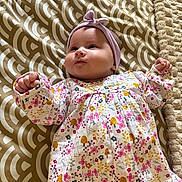 Léonie participe au concours pour gagner de l'argent avec cette photo : baby, infant, child, headband, floral_dress, blanket, pattern, cute, lying_down, indoors, soft_texture, raised_fists, cheeks, curious, young, adorable, portrait, clothing, headwear, person
