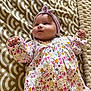 baby, infant, child, headband, floral_dress, blanket, pattern, cute, lying_down, indoors, soft_texture, raised_fists, cheeks, curious, young, adorable, portrait, clothing, headwear, person