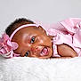 Anaiah is registered to the contest to win money with this photo: baby, infant, smiling, pink_dress, headband, flower, pearls, cute, happy, lying_down, fluffy_surface, portrait, child, closeup, skin, face, eyes, mouth, celebration, soft_texture