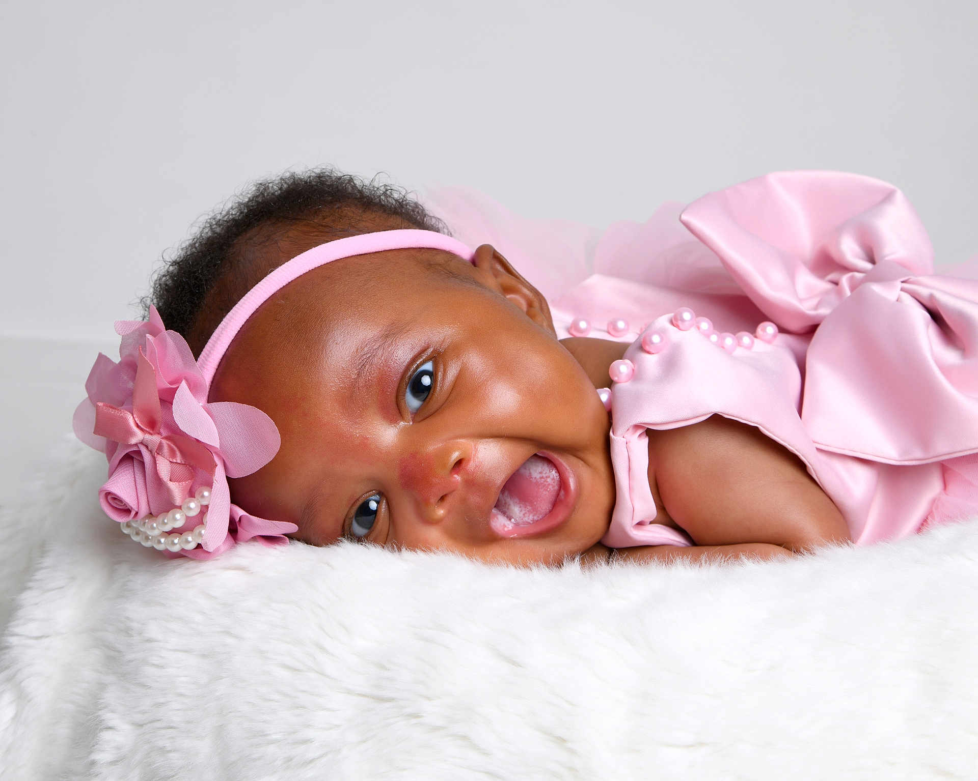 Anaiah is registered to the contest to win money with this photo: baby, infant, smiling, pink_dress, headband, flower, pearls, cute, happy, lying_down, fluffy_surface, portrait, child, closeup, skin, face, eyes, mouth, celebration, soft_texture