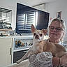cabinet, casual, chihuahua, dog, face, figurines, furniture, glasses, holding, home_decor, indoor, lace_blouse, light, living_room, pet, portrait, smile, television, woman, wooden_floor