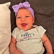 Cora is registered to the contest to win money with this photo: baby, infant, child, smiling, happy, portrait, close_up, purple_bow, headband, onesie, clothing, couch, pillow, indoor, sitting, lying_down, face, eyes, mouth, family_photo