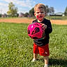 child, toddler, boy, soccer, football, soccer_ball, pink_ball, outdoor, sports_field, grass, sunny, blue_sky, smiling, portrait, standing, shorts, black_shirt, shoes, playground, baseball_field