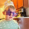 Wesley joined the competition — help win amazing prizes! child, toddler, portrait, face, glasses, star_shaped_glasses, paper_hat, hat, bib, patterned_clothing, kitchen, indoors, warm_lighting, cabinet, countertop, coffee_machine, seated, close_up, fashionable, playful