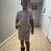 Cristopher is registered to the contest to win money with this photo: boy, casual, child, clothing, footwear, hallway, holding_object, indoor, patterned, person, portrait, serious, shirt, shoes, shorts, sneakers, standing, tile_floor, yellow_object, young