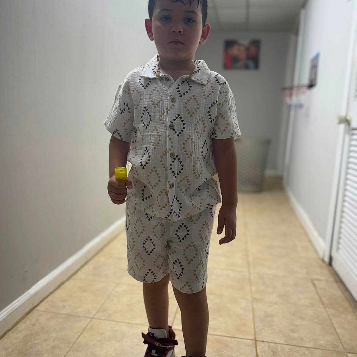 Cristopher is registered to the contest to win money with this photo: boy, casual, child, clothing, footwear, hallway, holding_object, indoor, patterned, person, portrait, serious, shirt, shoes, shorts, sneakers, standing, tile_floor, yellow_object, young
