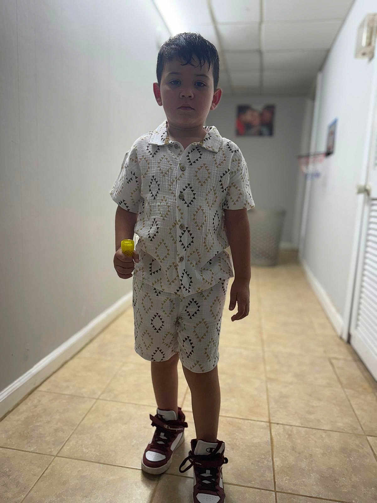 Cristopher is registered to the contest to win money with this photo: boy, casual, child, clothing, footwear, hallway, holding_object, indoor, patterned, person, portrait, serious, shirt, shoes, shorts, sneakers, standing, tile_floor, yellow_object, young