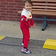 Matthew is registered to the contest to win money with this photo: child, toddler, smiling, walking, outdoor, portrait, red_tracksuit, sportswear, sneakers, curly_hair, bench, brick_wall, pavement, yellow_curb, playground, standing, young_person, casual_clothing, face, daylight
