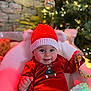 baby, child, red_hat, red_outfit, christmas_tree, holiday_decor, gold_ribbon, glowing_light, cozy_chair, smiling, festive, indoor, holiday, cute, celebration, happy, portrait, seasonal, knit_hat, decorations
