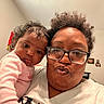 baby, infant, adult, parent, selfie, portrait, face, smiling, duck_face, eyeglasses, curly_hair, pink_clothing, indoor, family, closeup, shoulder, home, wall, photographs, cozy