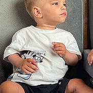 Lorenzo participe au concours pour gagner de l'argent avec cette photo : toddler, child, mickey_mouse, tshirt, black_shorts, sneakers, shoe, couch, gray_couch, seated, side_profile, short_hair, casual_clothing, indoors, young_child, looking_away, small_chip, hand_visible, light_skin, curious_expression