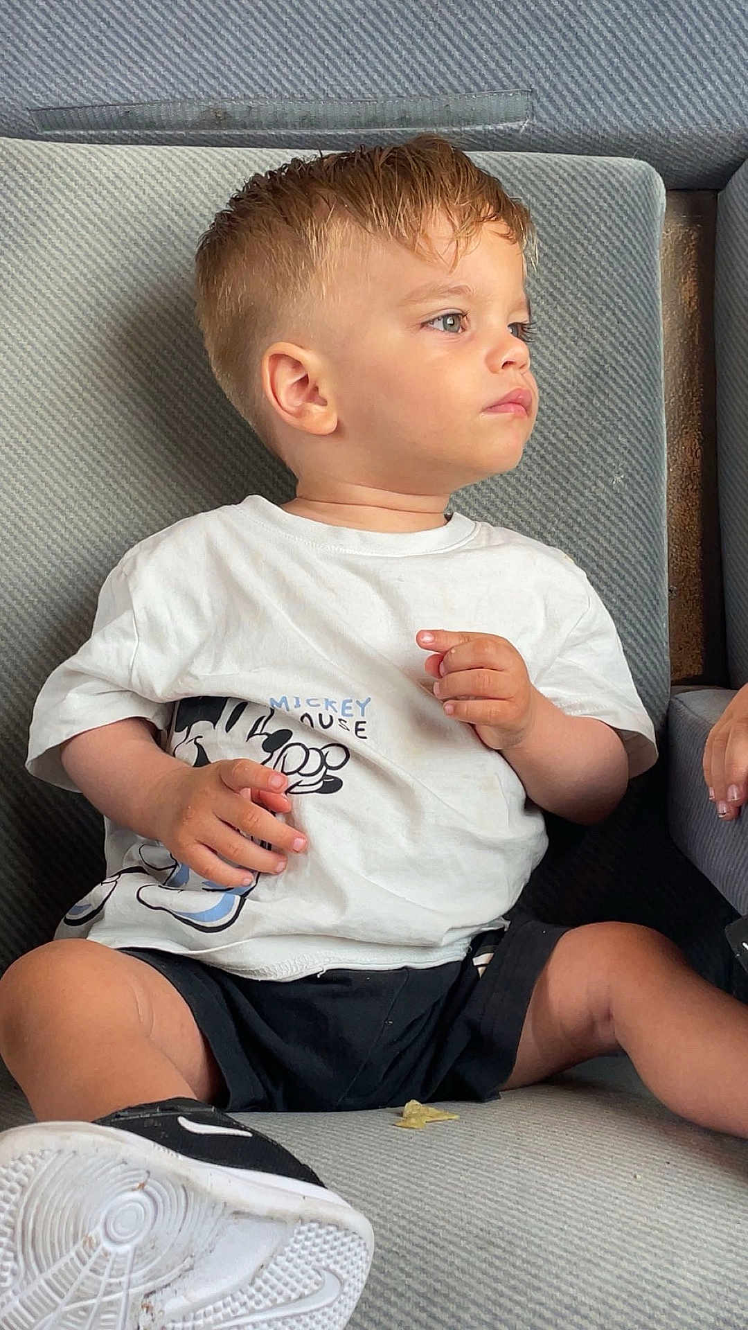 Lorenzo participe au concours pour gagner de l'argent avec cette photo : toddler, child, mickey_mouse, tshirt, black_shorts, sneakers, shoe, couch, gray_couch, seated, side_profile, short_hair, casual_clothing, indoors, young_child, looking_away, small_chip, hand_visible, light_skin, curious_expression